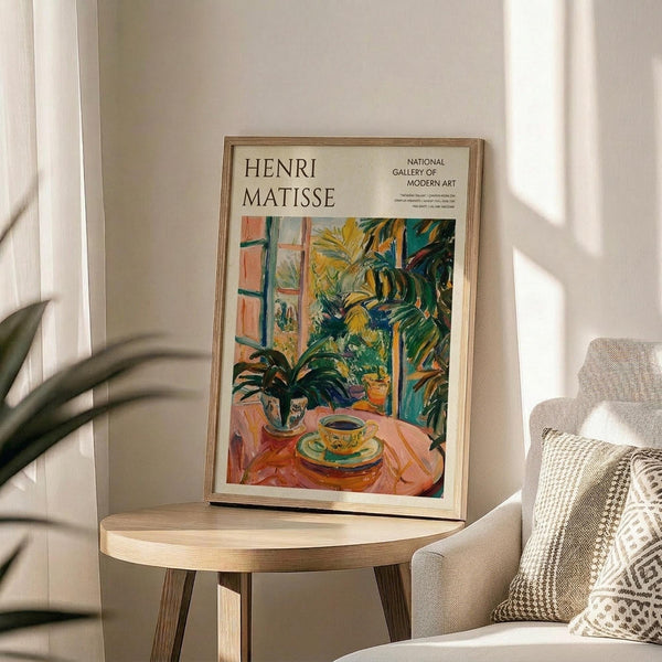 May include: Framed Henri Matisse museum exhibition poster featuring vibrant tropical interior scene with palm plants, teacup on round table, and bright greens, yellows, and oranges. Natural wood frame sits on light wooden side table next to beige upholstered chair with patterned throw pillows. Soft window light creates shadows on neutral wall.