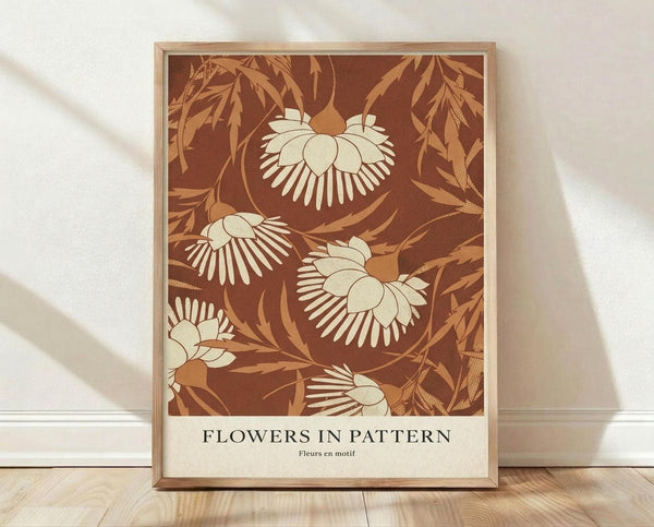 May include: Framed botanical art print with cream and brown floral pattern featuring stylized flowers with radiating petals and flowing leaves on rich brown background. Print displays "FLOWERS IN PATTERN" and "Fleurs en motif" text at bottom. Light wood frame leans against white textured wall on natural wood floor with soft window light casting shadows.