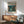 May include: Framed vintage-style poster showing a lone pilot in military gear walking through a rainy futuristic Tokyo street with towering mechs in the background, displayed above a mid-century modern wooden credenza with white ceramic lamp, next to a mint green upholstered armchair with wooden frame in a sunlit room with white walls.