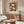 May include: Mid-century modern living room featuring a framed botanical poster with cream and rust-colored daisy-like flowers against a terracotta background. The print hangs above a walnut wood credenza with brass hardware. A white curved chair, brass floor lamp, glass-top coffee table, and neutral walls complete the warm, minimalist interior styling.