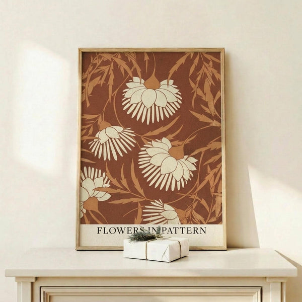 May include: Framed botanical art print with brown background featuring cream-colored stylized flowers and golden foliage in a decorative pattern. The poster displays "FLOWERS IN PATTERN" text at bottom and sits on a light wood console table beside a small wrapped gift with pine sprig. Natural lighting creates soft shadows on white wall behind the thin wooden frame.