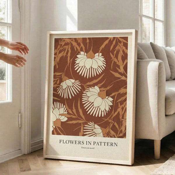 May include: Framed botanical art print with warm brown background featuring cream and orange tropical flowers and foliage in decorative pattern. White matted frame leans against beige armchair in bright living room with large windows and wooden floors. Person's hands gesture toward artwork.