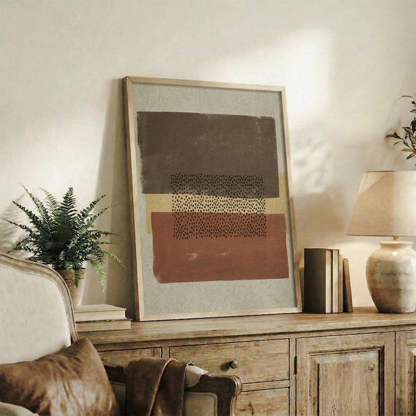 May include: Abstract geometric art print in light wooden frame displayed on rustic wooden sideboard. Features layered horizontal shapes in brown, terracotta, and cream tones with dotted texture details against light background. Styled with fern plant, ceramic vase, stacked books, and cream upholstered chair in warm-toned living room setting.