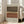 May include: Large framed abstract art print with horizontal bands in earth tones - sage green, dark brown, terracotta orange, and cream. Features scattered black dots across the middle sections creating geometric patterns. Light wood frame leans against white wall next to beige upholstered armchair in bright, airy living room with tall windows.
