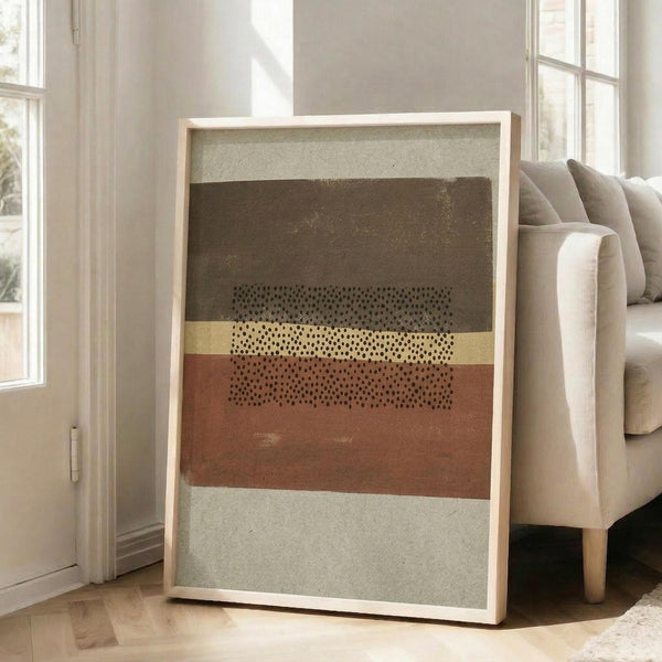 May include: Large framed abstract art print with horizontal bands in earth tones - sage green, dark brown, terracotta orange, and cream. Features scattered black dots across the middle sections creating geometric patterns. Light wood frame leans against white wall next to beige upholstered armchair in bright, airy living room with tall windows.