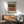 May include: Abstract geometric poster with earthy brown, rust orange, and cream horizontal bands featuring dotted pattern details, displayed in thin copper frame above mid-century modern walnut credenza. Sage green upholstered armchair with wooden arms sits beside round coffee table in bright, minimalist living room with white walls and natural sunlight creating geometric shadows.