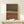 May include: Framed abstract art print with horizontal stripes in earth tones - dark brown at top, dotted pattern bands in gray and cream, solid rust orange at bottom. Light wood frame leaning against white wall on pale wooden surface. Small white wrapped gift box with evergreen sprig sits in front of artwork.