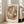 May include: Abstract geometric art print in warm earth tones featuring overlapping curved shapes in browns, tans, blues and greens, displayed in a light wood frame leaning against a beige sofa in a bright living room with white walls and large windows
