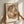 May include: Hands positioning a framed abstract art print on a white textured wall. The artwork features overlapping organic shapes in warm earth tones - terracotta, sage green, dusty pink, and blue - creating a layered, translucent effect. Natural light casts soft shadows across the scene. The print is displayed in a light wood frame with white matting.