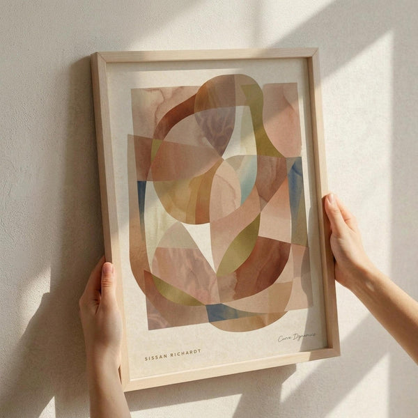 May include: Hands positioning a framed abstract art print on a white textured wall. The artwork features overlapping organic shapes in warm earth tones - terracotta, sage green, dusty pink, and blue - creating a layered, translucent effect. Natural light casts soft shadows across the scene. The print is displayed in a light wood frame with white matting.