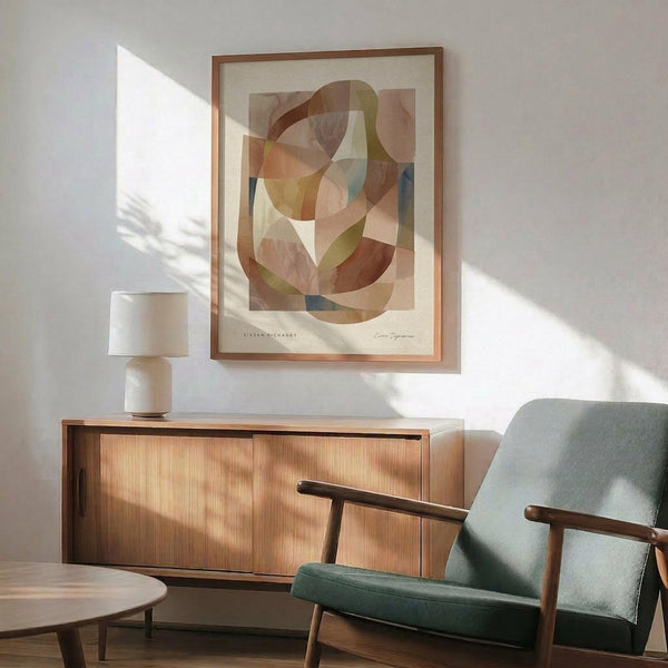 May include: Modern abstract art print with overlapping curved shapes in warm earth tones - browns, oranges, yellows, and blues - displayed in thin wooden frame above mid-century modern credenza. Sage green upholstered armchair with wooden arms sits nearby, white ceramic table lamp on credenza, natural sunlight casting shadows on white wall.