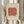 May include: Person holding a framed abstract art print featuring a coral-red painted rectangle with olive green dots on a cream background with pale blue-green vertical stripes. Light wood frame, styled with dried pampas grass against white curtains in soft natural lighting.