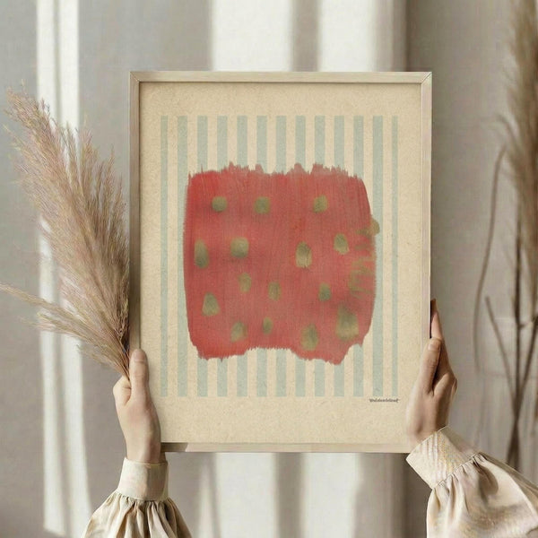 May include: Person holding a framed abstract art print featuring a coral-red painted rectangle with olive green dots on a cream background with pale blue-green vertical stripes. Light wood frame, styled with dried pampas grass against white curtains in soft natural lighting.
