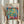 May include: Person in cream ruffled sleeves holding framed Henri Matisse poster featuring colorful open window scene with tropical plants, teacup on pink table, vibrant blues, greens, oranges. Dried pampas grass visible left side. Light wooden frame against soft neutral curtain background.