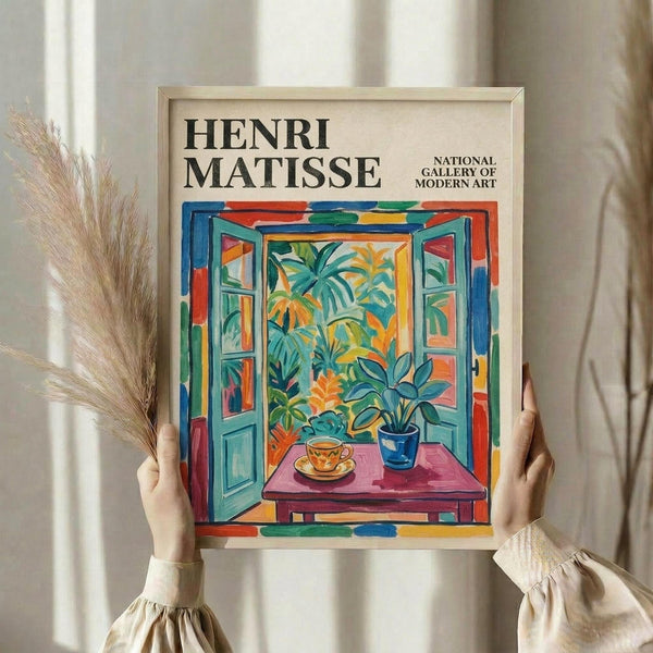 May include: Person in cream ruffled sleeves holding framed Henri Matisse poster featuring colorful open window scene with tropical plants, teacup on pink table, vibrant blues, greens, oranges. Dried pampas grass visible left side. Light wooden frame against soft neutral curtain background.