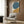 May include: Abstract poster with large teal blue oval and mustard yellow curved shape on cream background, displayed in light wood frame on textured wood wall above beige upholstered armchair with geometric patterned pillow, brass floor lamp and dark side table with open book nearby