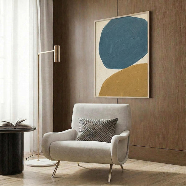May include: Abstract poster with large teal blue oval and mustard yellow curved shape on cream background, displayed in light wood frame on textured wood wall above beige upholstered armchair with geometric patterned pillow, brass floor lamp and dark side table with open book nearby