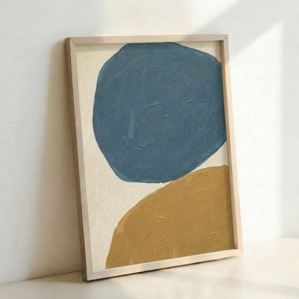 Abstract painting in light wood frame featuring large teal blue circular shape in upper portion and golden mustard yellow curved form in lower right corner against cream textured background. Visible brushstrokes create organic, hand-painted appearance. Frame leans against white wall casting soft shadow.