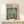 May include: Framed Henri Matisse exhibition poster featuring colorful open window scene with tropical plants, teacup on pink table, and vibrant blue shutters. Thin wooden frame displayed on light wooden shelf beside white wrapped gift with greenery sprig against cream wall.