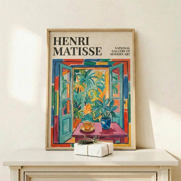 May include: Framed Henri Matisse exhibition poster featuring colorful open window scene with tropical plants, teacup on pink table, and vibrant blue shutters. Thin wooden frame displayed on light wooden shelf beside white wrapped gift with greenery sprig against cream wall.