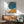 May include: Abstract art print with large teal blue circle and golden yellow curved shape on cream background, displayed in thin wooden frame above mid-century modern walnut credenza. Scene features sage green upholstered armchair with wood arms, white ceramic table lamp, and soft natural lighting casting shadows on white wall.