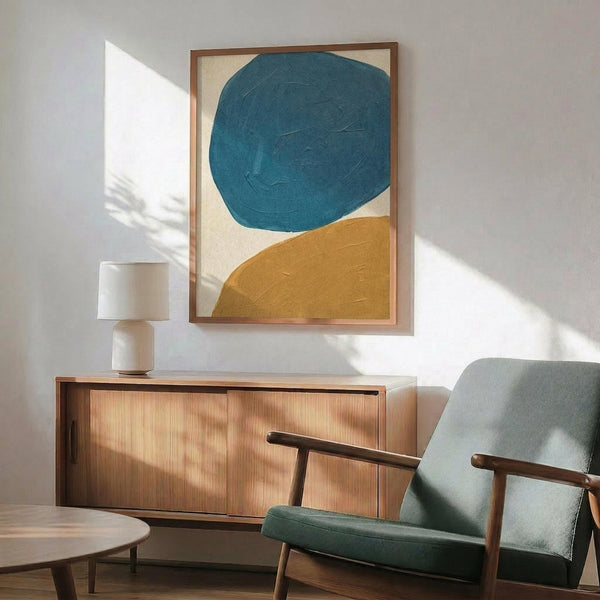 May include: Abstract art print with large teal blue circle and golden yellow curved shape on cream background, displayed in thin wooden frame above mid-century modern walnut credenza. Scene features sage green upholstered armchair with wood arms, white ceramic table lamp, and soft natural lighting casting shadows on white wall.