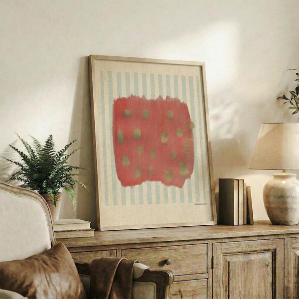 May include: Framed abstract art print with coral red brushstroke shape containing sage green dots over light blue and cream vertical stripes, displayed on rustic wooden sideboard alongside fern plant, ceramic vases, and books in neutral-toned living room setting.