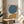May include: Abstract minimalist artwork in thin wood frame displaying teal blue circular shape and mustard yellow curved form on cream background, positioned on round wooden side table next to beige upholstered chair with geometric patterned throw pillow, natural lighting creating soft shadows on neutral wall with leafy plant visible in corner