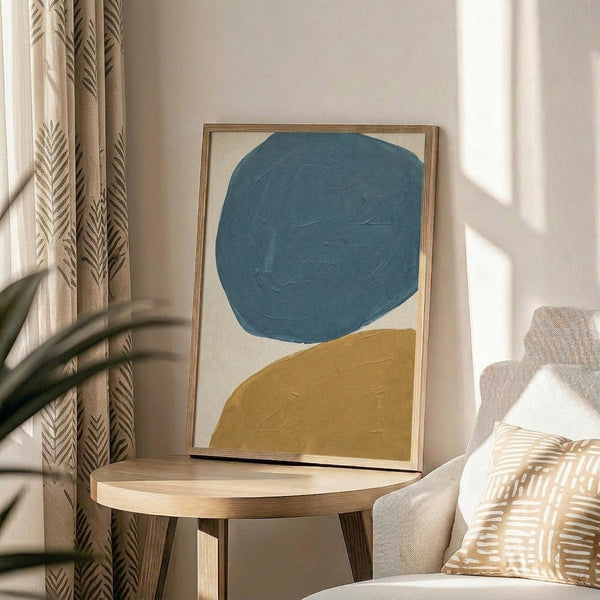 May include: Abstract minimalist artwork in thin wood frame displaying teal blue circular shape and mustard yellow curved form on cream background, positioned on round wooden side table next to beige upholstered chair with geometric patterned throw pillow, natural lighting creating soft shadows on neutral wall with leafy plant visible in corner