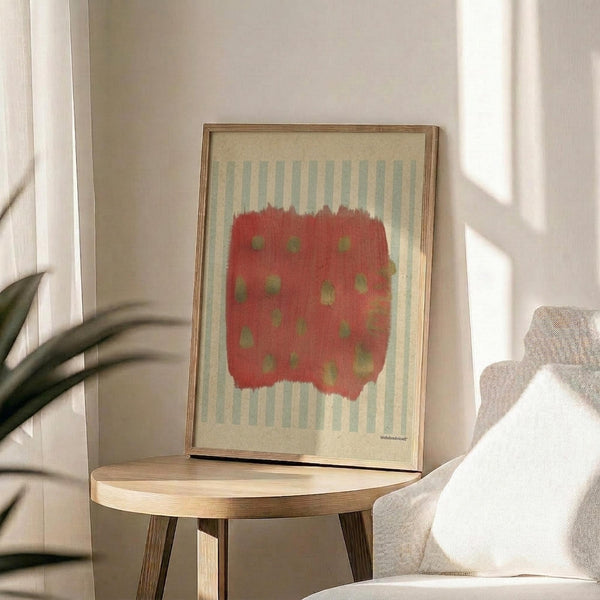 May include: Abstract art print in wooden frame sitting on round wooden side table next to beige upholstered chair. Artwork features red brushstroke shape with green dots against cream background with vertical blue-gray stripes. Soft natural lighting creates shadows on white wall. Plant leaves visible in corner.