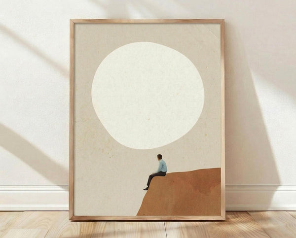 May include: Minimalist art print in light wood frame leaning against white textured wall on light wooden floor. Artwork features small figure in light blue shirt sitting on terracotta curved hill beneath large cream circle resembling moon against beige background. Soft natural lighting creates gentle shadows on wall.