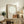 May include: Minimalist poster artwork displayed in wooden frame on rustic sideboard, featuring small figure sitting on terracotta hillside beneath large cream circle against beige background. Room setting shows leather armchair, fern plant, stacked books, ceramic vase, and soft natural lighting creating warm, contemplative atmosphere.