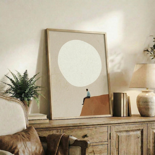 May include: Minimalist poster artwork displayed in wooden frame on rustic sideboard, featuring small figure sitting on terracotta hillside beneath large cream circle against beige background. Room setting shows leather armchair, fern plant, stacked books, ceramic vase, and soft natural lighting creating warm, contemplative atmosphere.