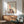 May include: Colorful abstract art print in wooden frame showing stylized figure with red hair reaching toward a star, lower body formed by geometric houses in blues, reds, and yellows. Print displayed above mid-century wooden credenza with ceramic lamp in bright living room with sage green armchair, round coffee table, and natural sunlight streaming across white walls.