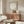 May include: Minimalist poster with beige background and large white circle, featuring small figure sitting on brown curved shape below. Artwork displayed in thin wood frame above walnut credenza with white mushroom lamp. Modern living room setting with cream walls, white sculptural chair, glass coffee table, and natural lighting creating warm shadows.