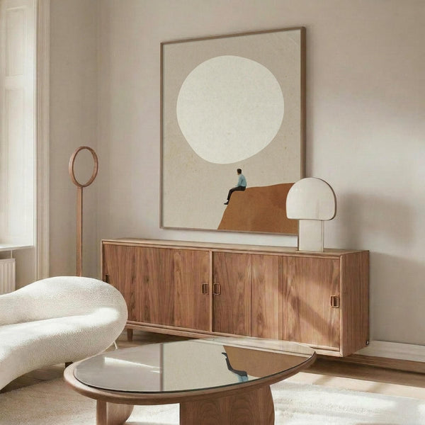 May include: Minimalist poster with beige background and large white circle, featuring small figure sitting on brown curved shape below. Artwork displayed in thin wood frame above walnut credenza with white mushroom lamp. Modern living room setting with cream walls, white sculptural chair, glass coffee table, and natural lighting creating warm shadows.