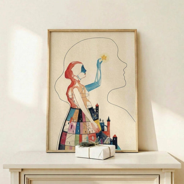 May include: Framed artistic poster showing a woman with red hair reaching toward a yellow star, her dress filled with colorful houses and buildings in a whimsical illustrated style. The artwork is displayed in a light wood frame on a white shelf next to a small wrapped gift, set against a cream-colored wall with soft natural lighting.