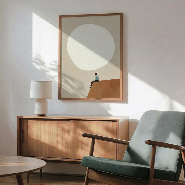 May include: Minimalist poster in wooden frame showing small figure sitting on orange-brown hill beneath large pale circle against beige background, displayed above wood sideboard with white table lamp, next to mid-century modern teal upholstered armchair with wooden arms, natural sunlight casting geometric shadows across white wall