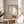 May include: Minimalist art print in wooden frame showing small figure sitting on geometric orange shape beneath large white circle on beige background, displayed on round wooden side table next to beige upholstered chair with plant leaves visible, natural lighting creating soft shadows on neutral wall