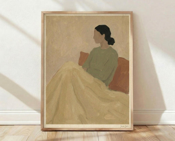 May include: Framed art print leaning against white wall on light wooden floor with natural shadows. Shows minimalist illustration of woman in profile sitting on orange-brown chair, wearing sage green top and flowing cream-colored skirt or blanket. Dark hair in low bun, faceless figure rendered in muted earth tones against beige background. Light wood frame with white matting.