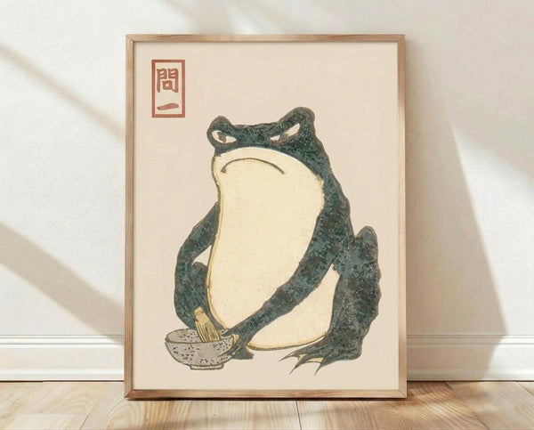 May include: Framed Japanese woodblock print featuring a green frog sitting upright with cream-colored belly, appearing contemplative with downturned mouth. The frog holds a small speckled bowl. Red seal stamp with Japanese characters appears in upper left corner. Artwork displayed in light wooden frame against white textured wall with soft shadows, sitting on light wood flooring.