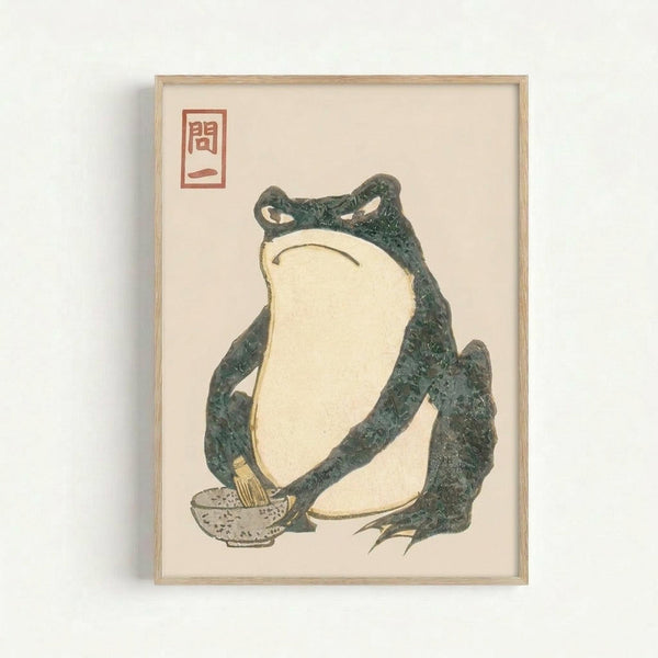 Framed vintage Japanese woodblock print featuring a large green frog with cream-colored belly sitting in a contemplative pose. The frog has detailed textured skin and is positioned next to what appears to be a small bowl or vessel. Red Japanese characters appear in the upper left corner against the cream background. The artwork is displayed in a simple light wood frame against a white wall.