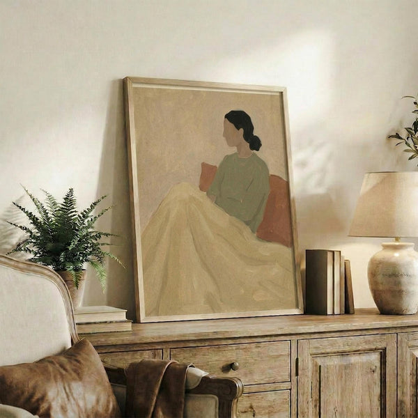 May include: Framed artwork showing a woman with dark hair in a low bun, wearing a sage green top and flowing yellow skirt, sitting against terracotta cushions in minimalist style with muted beige background. Print displayed on rustic wooden sideboard alongside fern plant, ceramic vases, books, and table lamp in warm-toned living room setting.