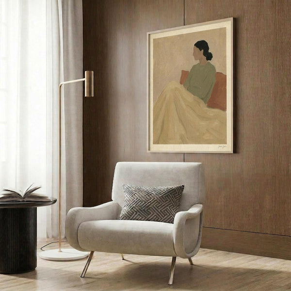 May include: Minimalist poster print featuring a seated woman with dark hair in a green top and flowing yellow skirt against a beige background, displayed in light wood frame on textured brown wood paneling. Modern reading corner with cream upholstered chair, geometric patterned pillow, brass floor lamp, and dark round side table with open book.