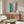 May include: Abstract art print with blue and pink rectangular color blocks crossed by dark blue brushstrokes, displayed in thin frame above mid-century walnut credenza. Modern living room setting with cream textured chair, brass floor lamp, oval glass coffee table, and soft natural lighting on neutral walls.