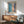 May include: Abstract artwork with blue and coral pink brushstrokes framed in natural wood, displayed above a mid-century modern walnut credenza with a ceramic table lamp. Sage green upholstered armchair with wooden frame sits nearby on hardwood floors. Soft natural light creates shadows on white wall in minimalist living room setting.