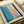 Abstract minimalist artwork featuring soft blue and pink rectangular color blocks with bold navy blue brushstrokes forming a loose frame around the composition. The piece has a watercolor-like texture with gentle color gradients and is displayed on cream-colored paper within an open art portfolio or sketchbook, surrounded by neutral-toned materials and ribbon.
