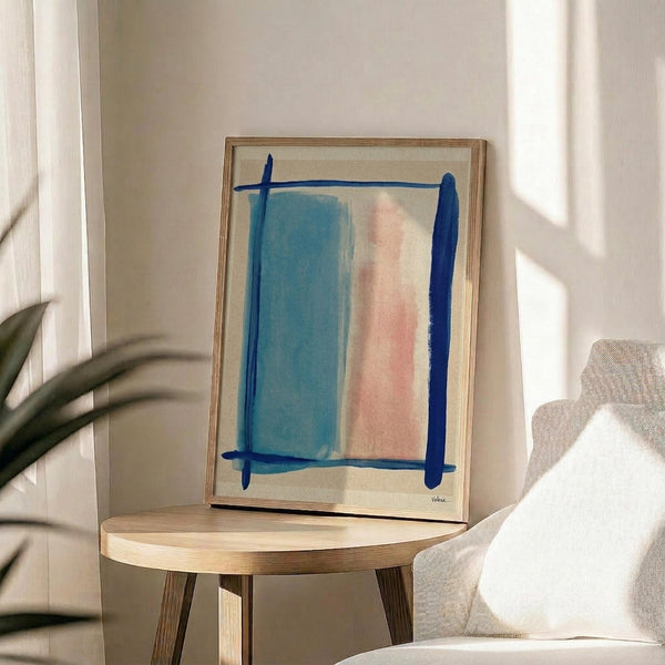 May include: Abstract minimalist artwork in light wood frame displayed on round wooden side table next to beige upholstered chair. The painting features soft blue and coral pink rectangular brushstrokes framed by dark blue painted lines on cream background. Natural sunlight creates shadows across white wall, with partial view of plant leaves in corner.