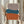 May include: Hands holding a framed abstract art print featuring three stacked organic shapes in muted terracotta orange, teal blue, and dusty mauve against a cream background. Natural light wooden frame. Person wearing beige ruffled sleeves holds dried pampas grass. Soft neutral curtained background creates minimalist styling.