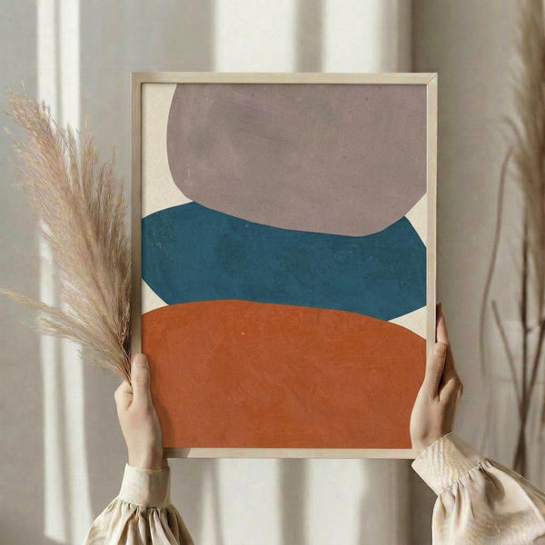 May include: Hands holding a framed abstract art print featuring three stacked organic shapes in muted terracotta orange, teal blue, and dusty mauve against a cream background. Natural light wooden frame. Person wearing beige ruffled sleeves holds dried pampas grass. Soft neutral curtained background creates minimalist styling.